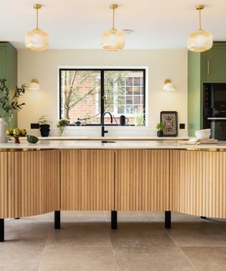 A kitchen with a light wooden kitchen island with a white counter, three gold pendant lights, a gray concrete tiled floor, and a black window