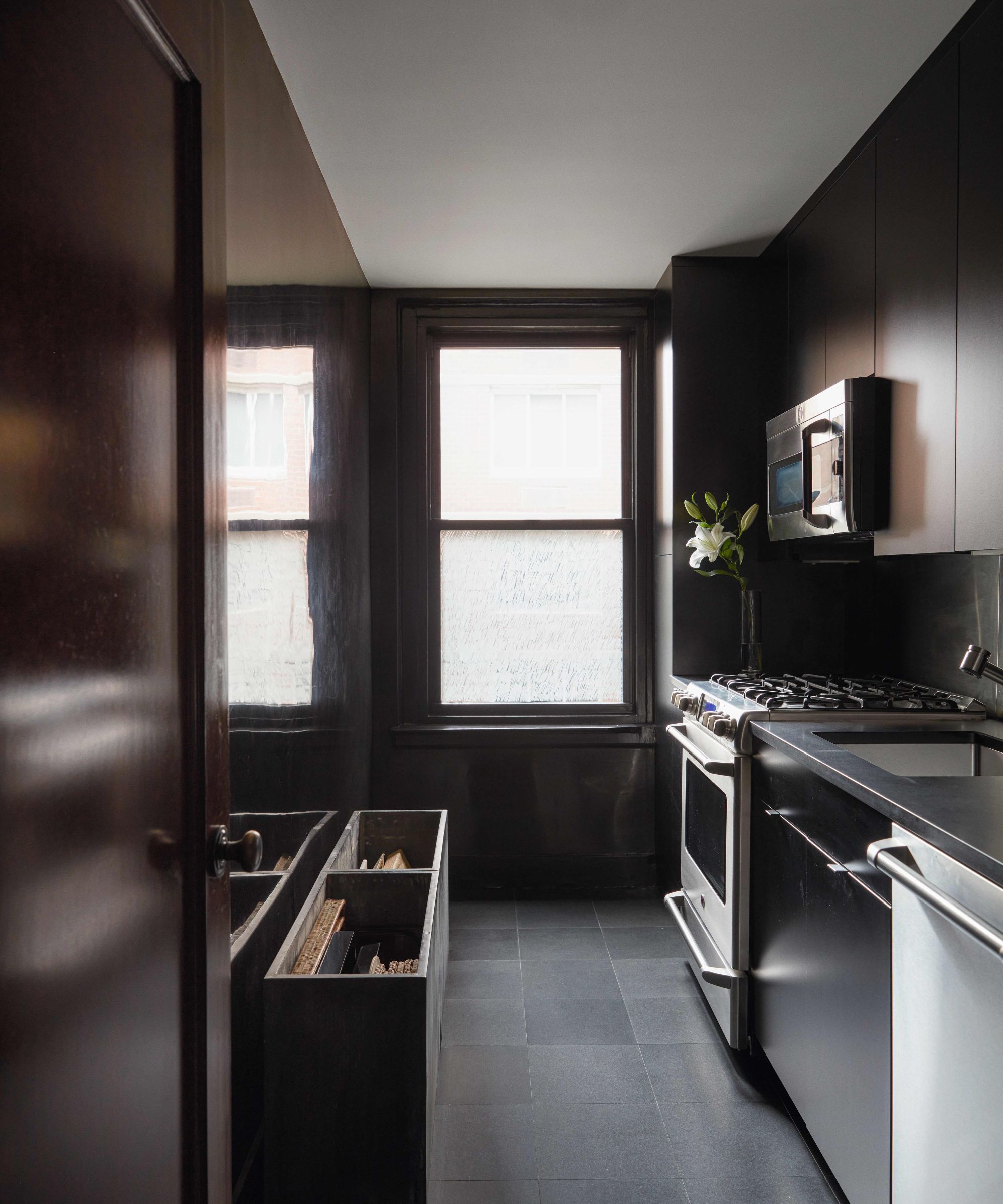 A narrow, modern galley kitchen with dramatic black cabinetry, a stainless steel range, and dark floor tiles, illuminated by soft light from a single window