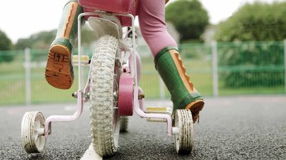 child with green dinosaur boots and pink pants on bike with training wheels