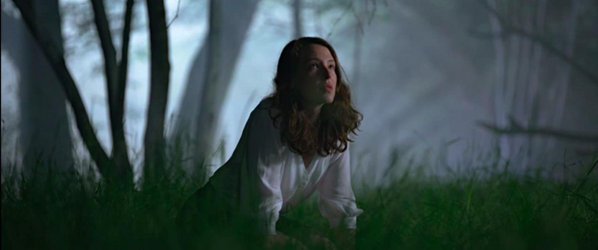 A young woman kneels in the grass in a deep forest looking enchanted