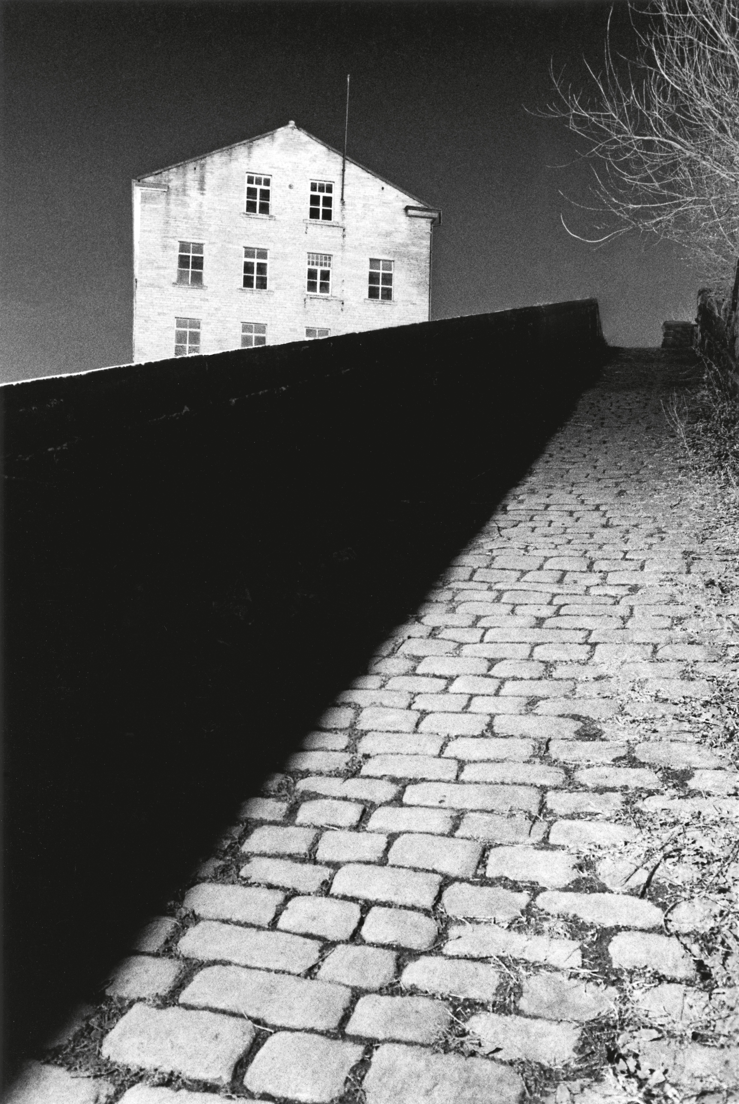 A steep, cobblestone path leads upward toward a multi-story industrial building under a dark, high-contrast sky.