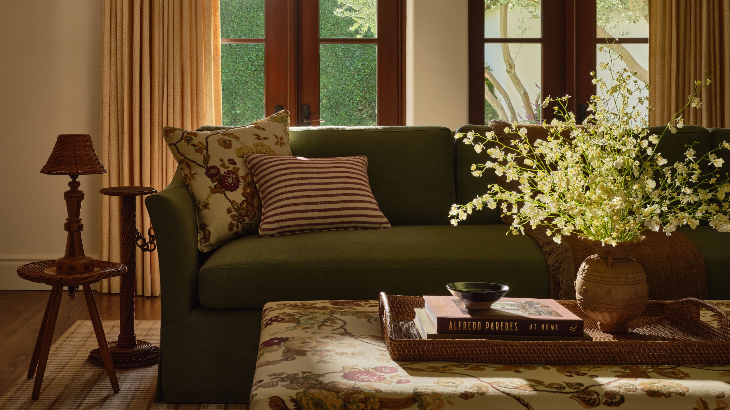 a cozy living room with dark wooden french doors onto the patio, a muted green sofa and floral upholstered pillows and matching ottoman