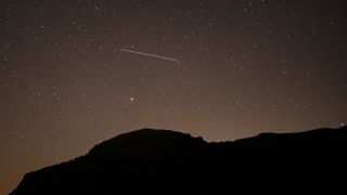A lone meteor is spotted streaking through a dark starry sky above a silhouetted hill.