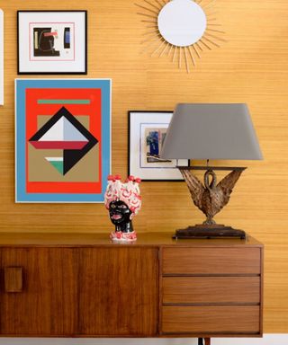 A bright yellow wall with a wooden sideboard, lamp and abstract red painting
