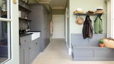 Pastel green utility room and mudroom gray cabinets, storage bench and butler sink. It has cream stone floors and mounted shelves and hooks for added storage.