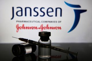 An image of two vials and a syringe in front of Johnson and Johnson&#039;s Janssen pharmaceutical companies sign.