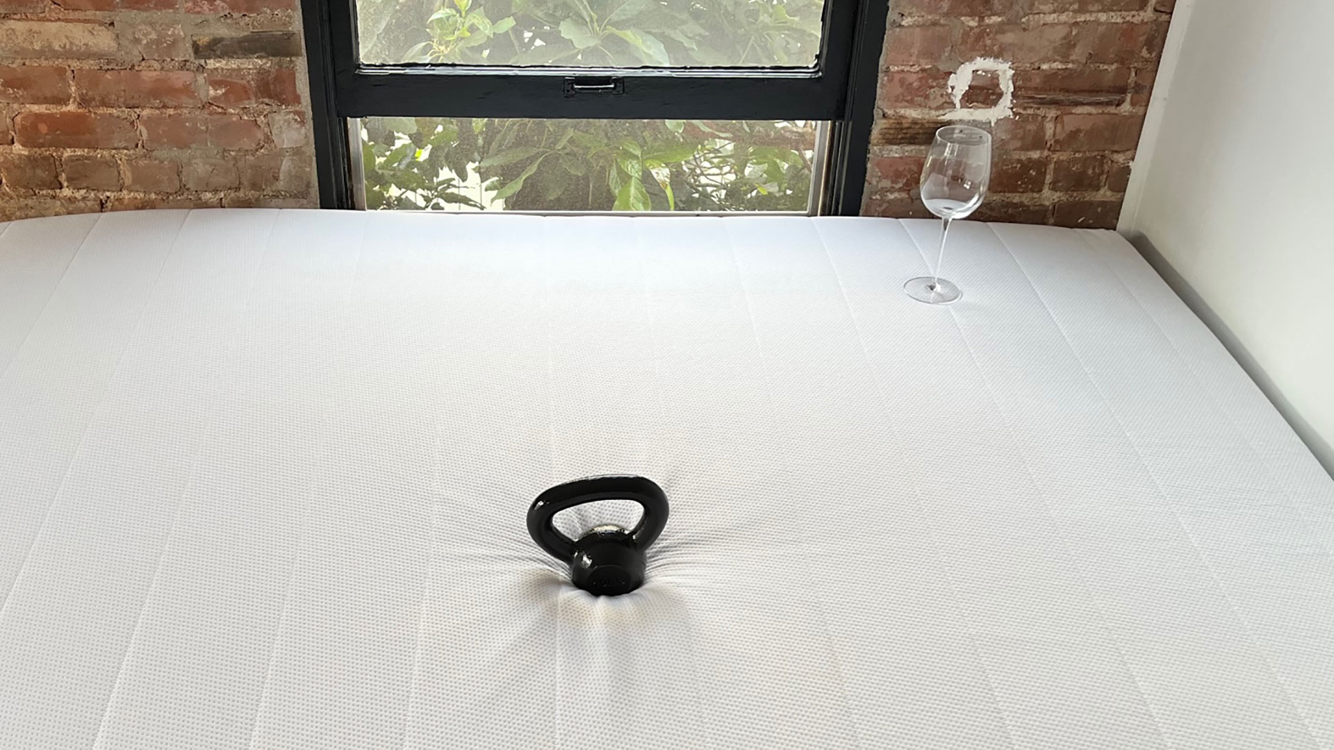 Photo of Helix Midnight mattress in a bedroom, with a weight on it, and a wine glass stood upright on the mattress surface nearby