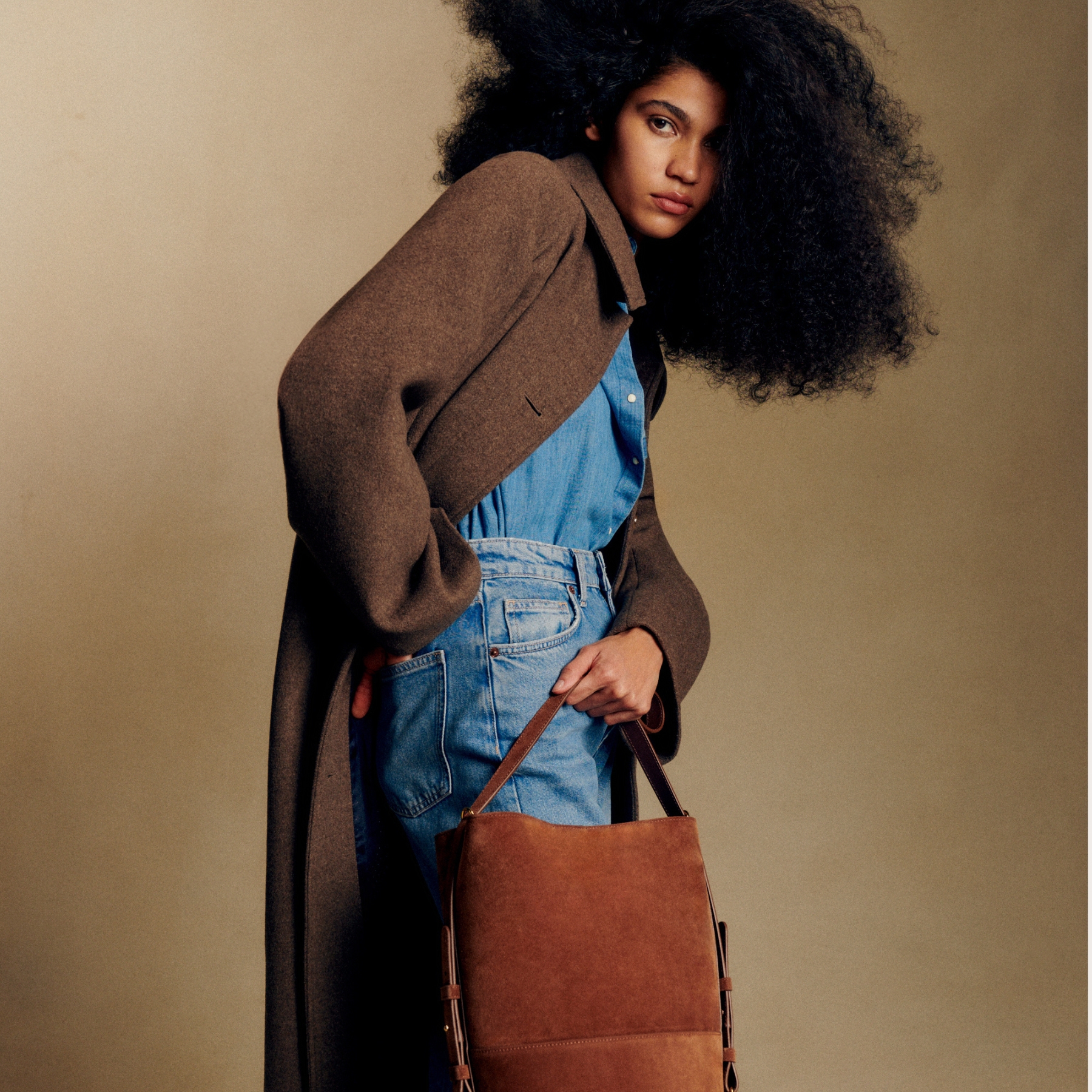 A model wearing a brown coat, blue shirt and jeans, with a suede bucket tote handbag.