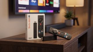 The new Amazon Fire TV Stick resting on a TV stand with a TV in the background showing the app home interface.