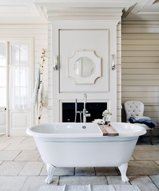 A white bathroom with a roll-top bath in the center, stone flooring and a fireplace behind