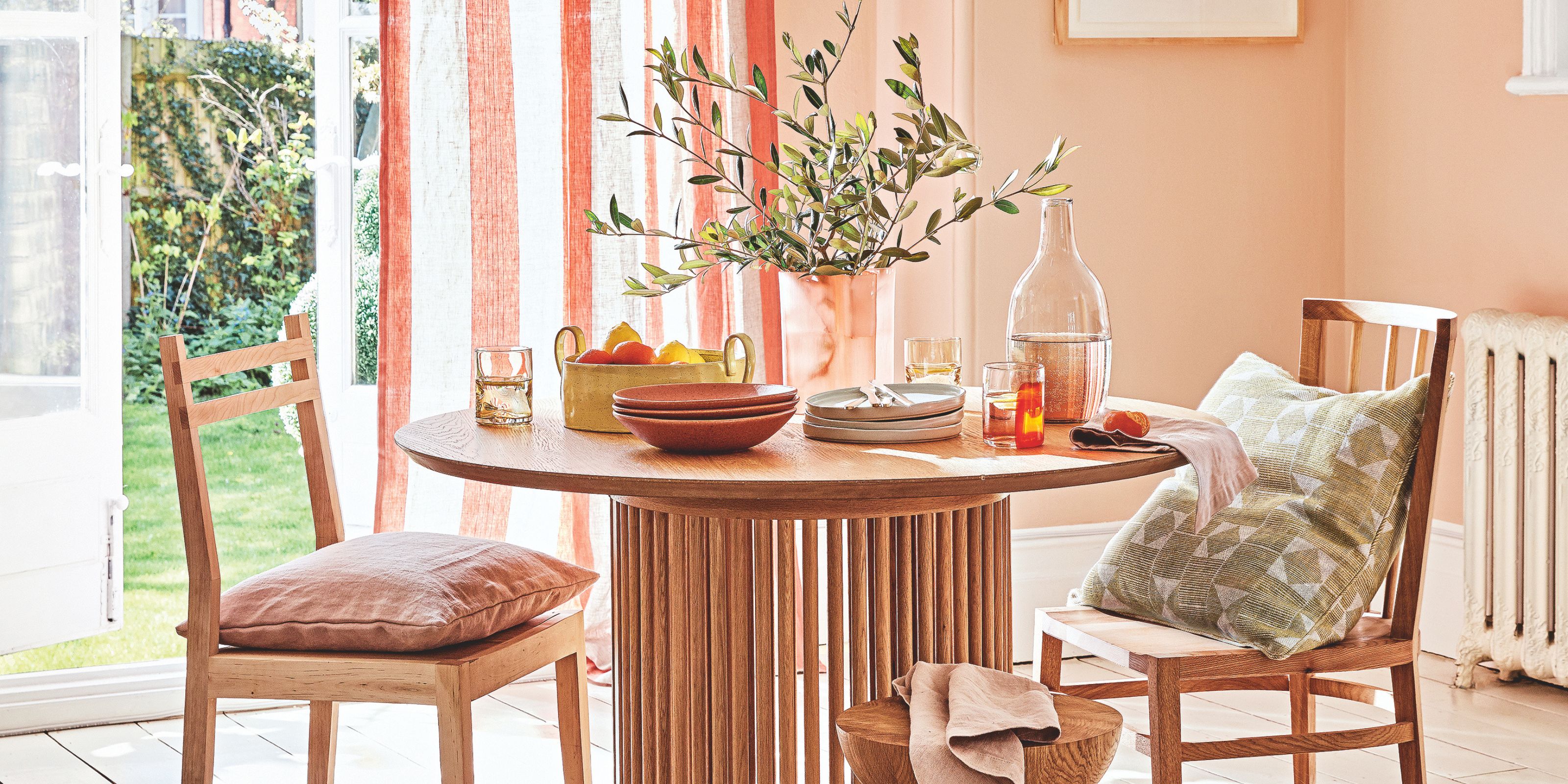A wooden table with bowls, plates and a fruit bowl on top within a pale pink room