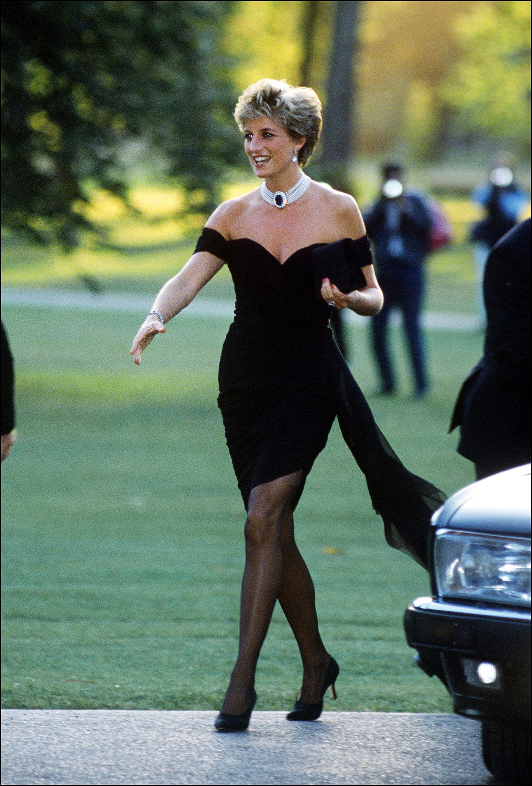 Princess Diana wearing a low-cut black dress walking next to a car