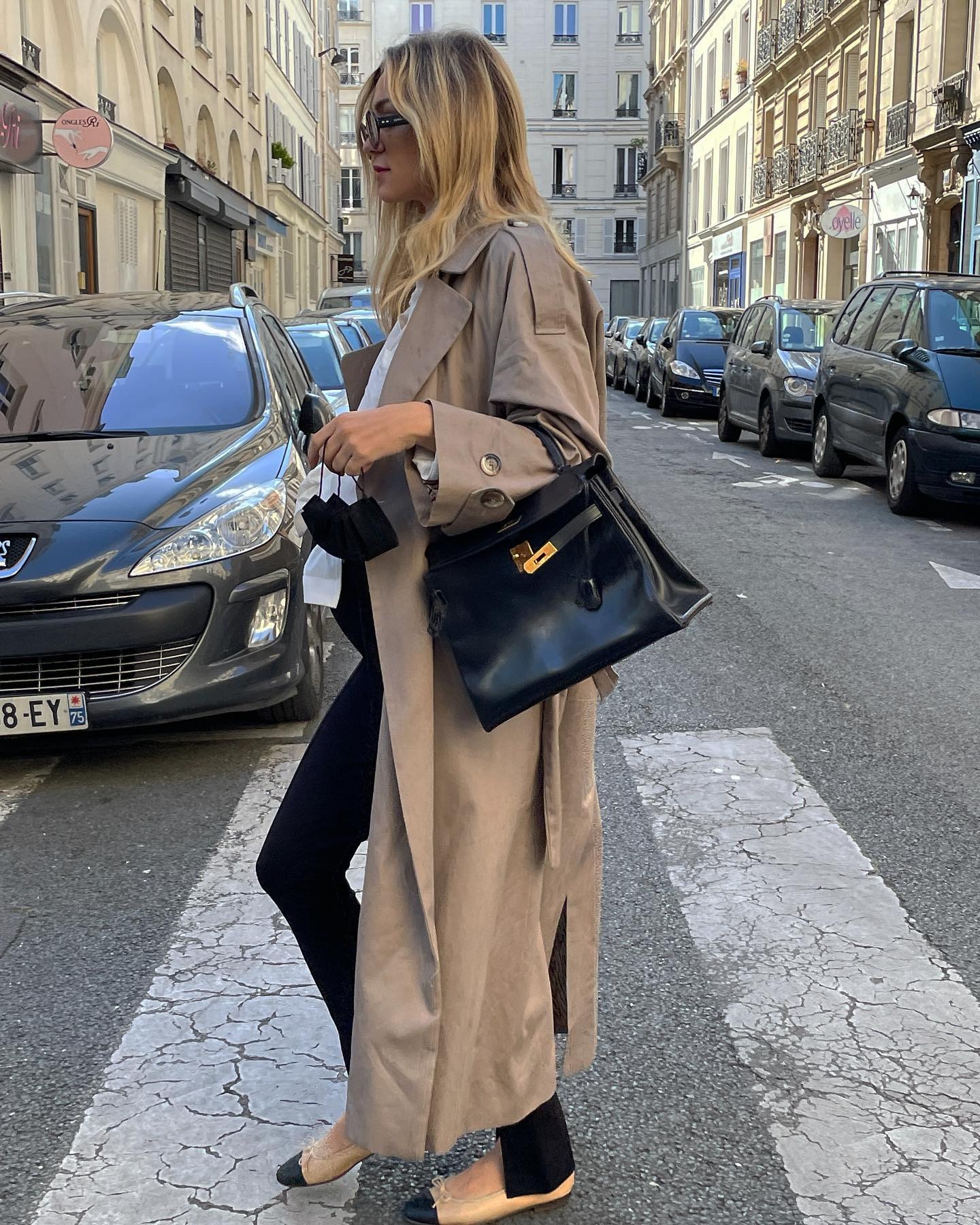 French influencer Anne-Laure Mais walks across a Paris street wearing a chic maternity outfit with sunglasses, a long trench coat, white shirt, black ankle-slit leggings, black Hermes bag, and two-tone Chanel ballet flats