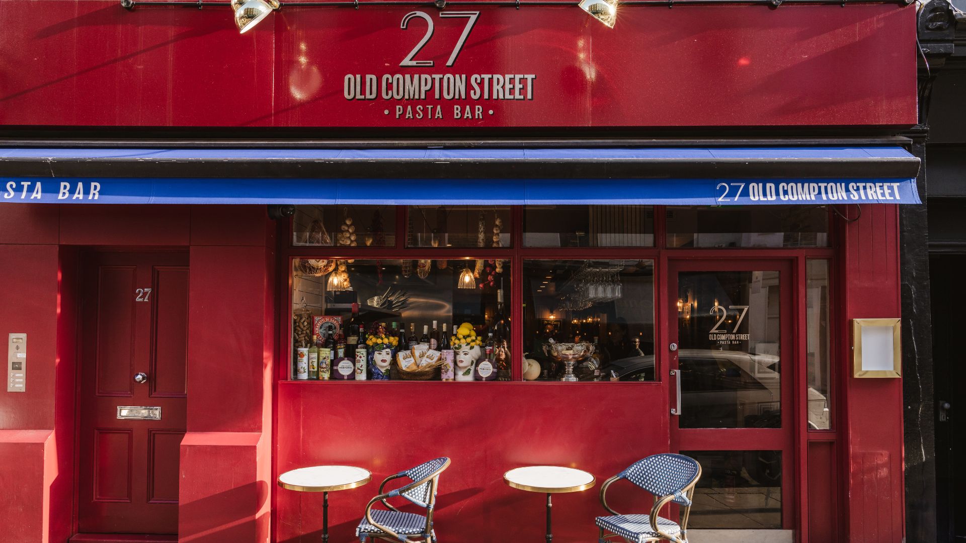 an image of the restaurant's exterior. It is a small, red building with its name written across the top in gold. It has two small round tables outside and blue and white wicker chairs next to them
