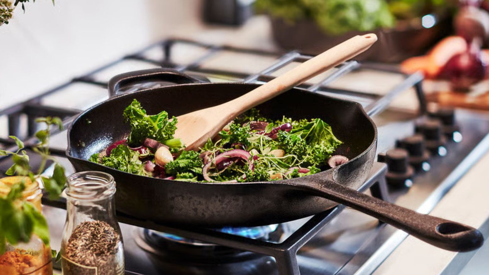 A black cast iron skillet on a gas stove, filled with green leafy vegetables and red onion. Surrounded by cooking ingredients, out of focus. 