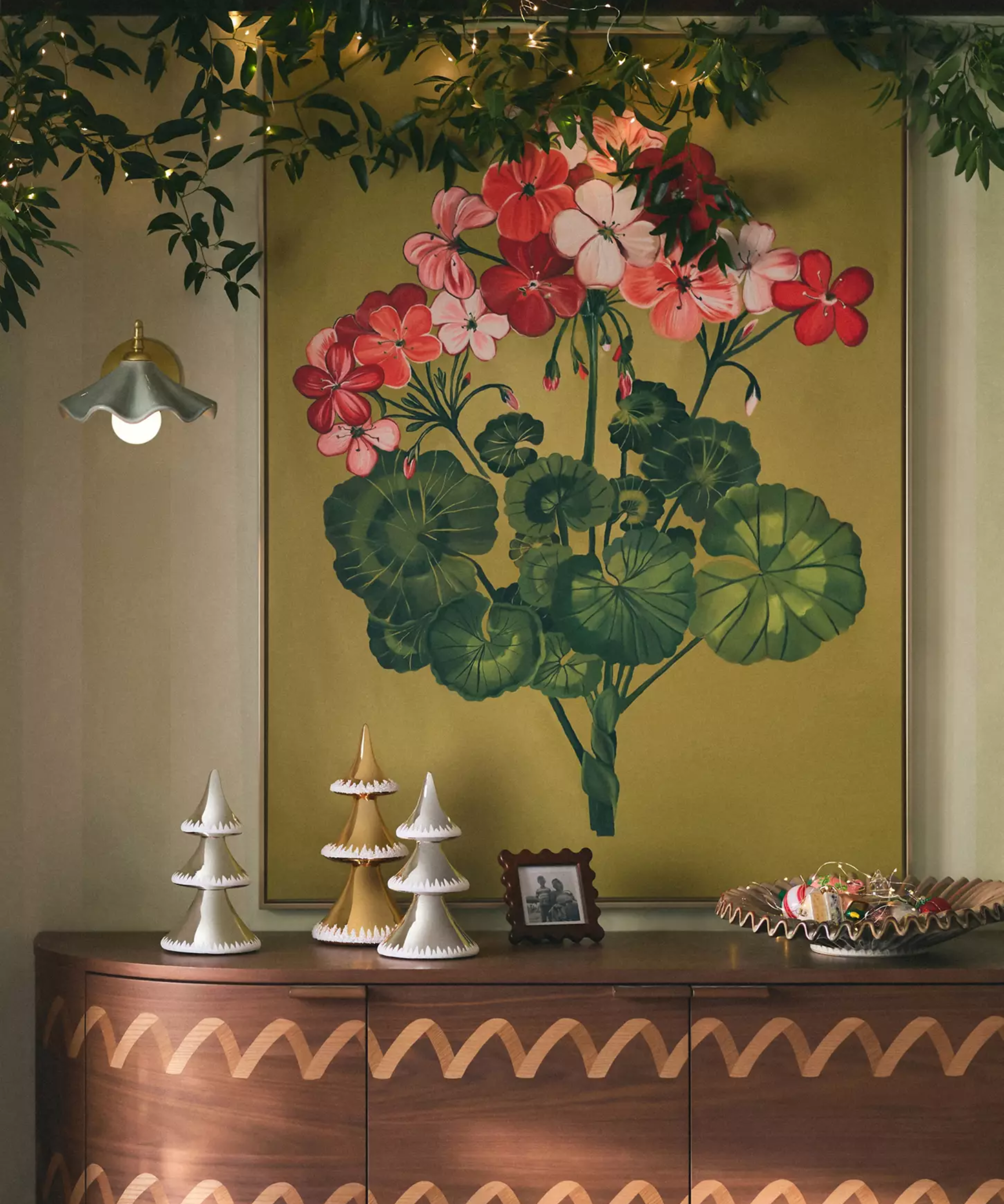 Festive credenza vignette featuring a metallic tree trio, pleated gold bowl, scalloped picture frame, playful ceramic wall sconce, oversized metallic art, and strings of warmly lit garland overhead