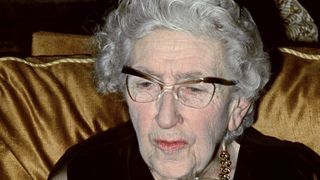 Agatha Christie in a black dress and glasses sits on a gold chair.