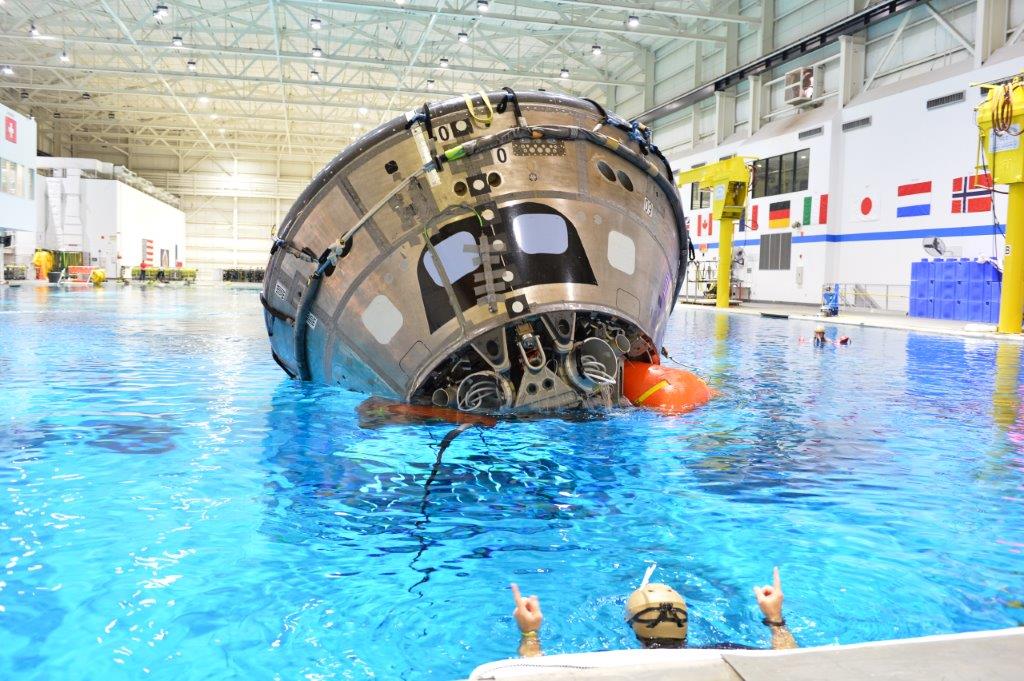 an inverted spacecraft capsule prototype floats in a pool with the help of submerged orange inflatable balloons. a diver near the front of the picture gestures up with his two index fingers. at far right is a wall of the neutral buoyancy laboratory with flags of different nations participating in training there