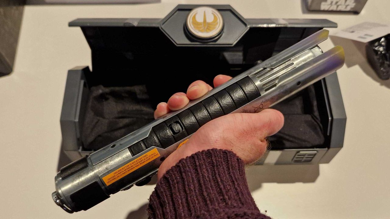 A hand holding the Master Sol lightsaber hilt, with an open case in the background