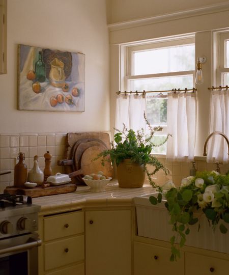 Sunlit rustic kitchen counter with cream tiles, potted herbs, fresh flowers in an apron sink, and a still-life painting.