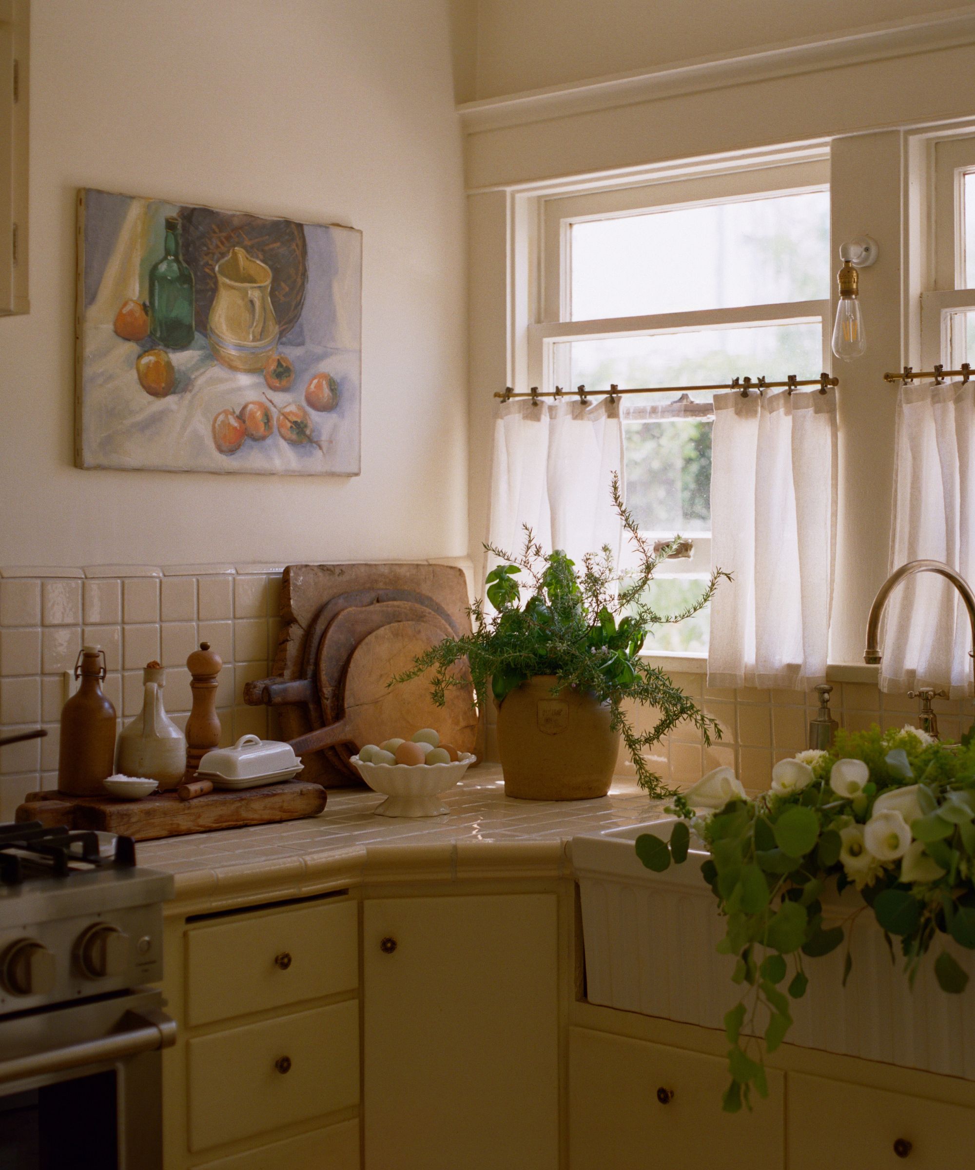 Sunlit rustic kitchen counter with cream tiles, potted herbs, fresh flowers in an apron sink, and a still-life painting.