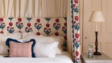 A horizontal shot of a bed set against a neutral paneled wall. The headboard and draped canopy curtains feature a vertical pattern of stylized red flowers with blue stems on a cream background. The bed has white ruffled shams and a small rectangular pillow with red dots and a thick navy blue ruffle. To the right, a white side table holds a clear floral-patterned carafe and a classic black and gold lamp with a cream shade.
