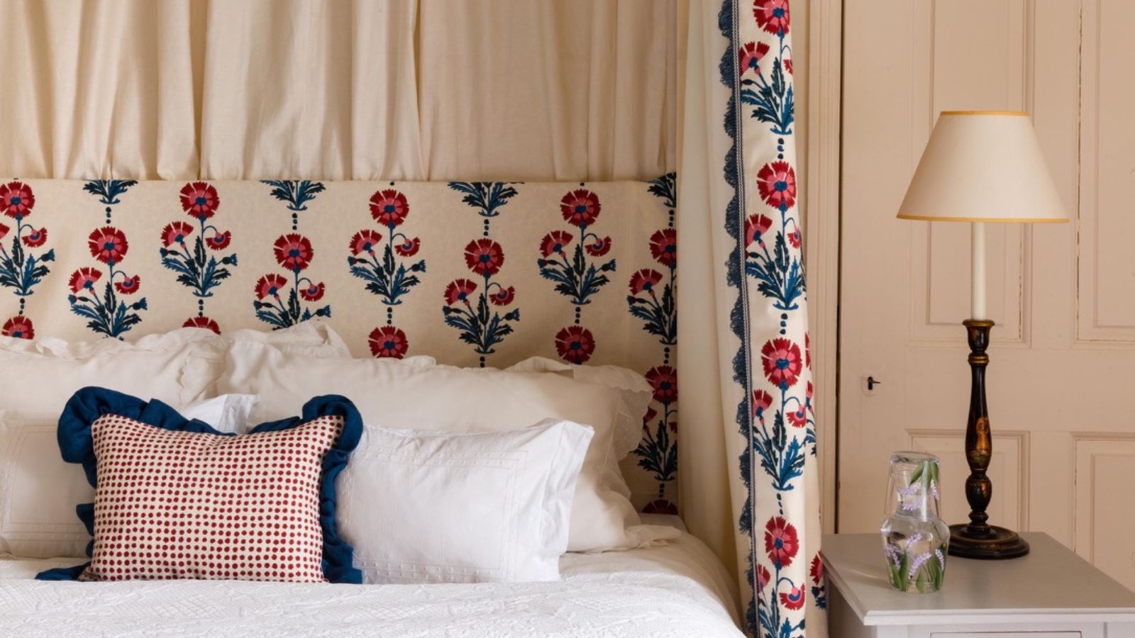 A horizontal shot of a bed set against a neutral paneled wall. The headboard and draped canopy curtains feature a vertical pattern of stylized red flowers with blue stems on a cream background. The bed has white ruffled shams and a small rectangular pillow with red dots and a thick navy blue ruffle. To the right, a white side table holds a clear floral-patterned carafe and a classic black and gold lamp with a cream shade.