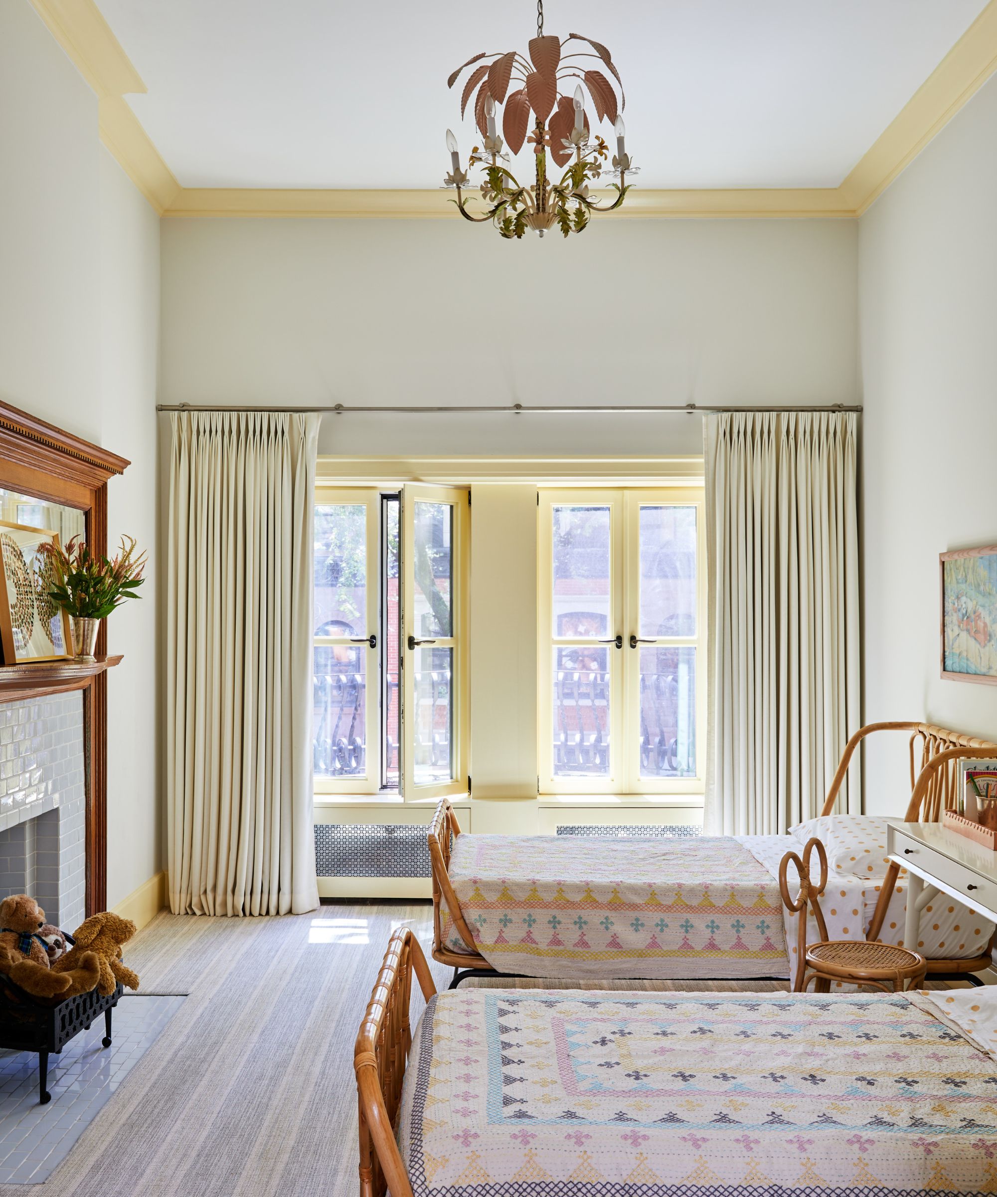 Tall, white bedroom with twin rattan beds, pale yellow window trim, a tiled fireplace with teddy bears, and a delicate wood chandelier