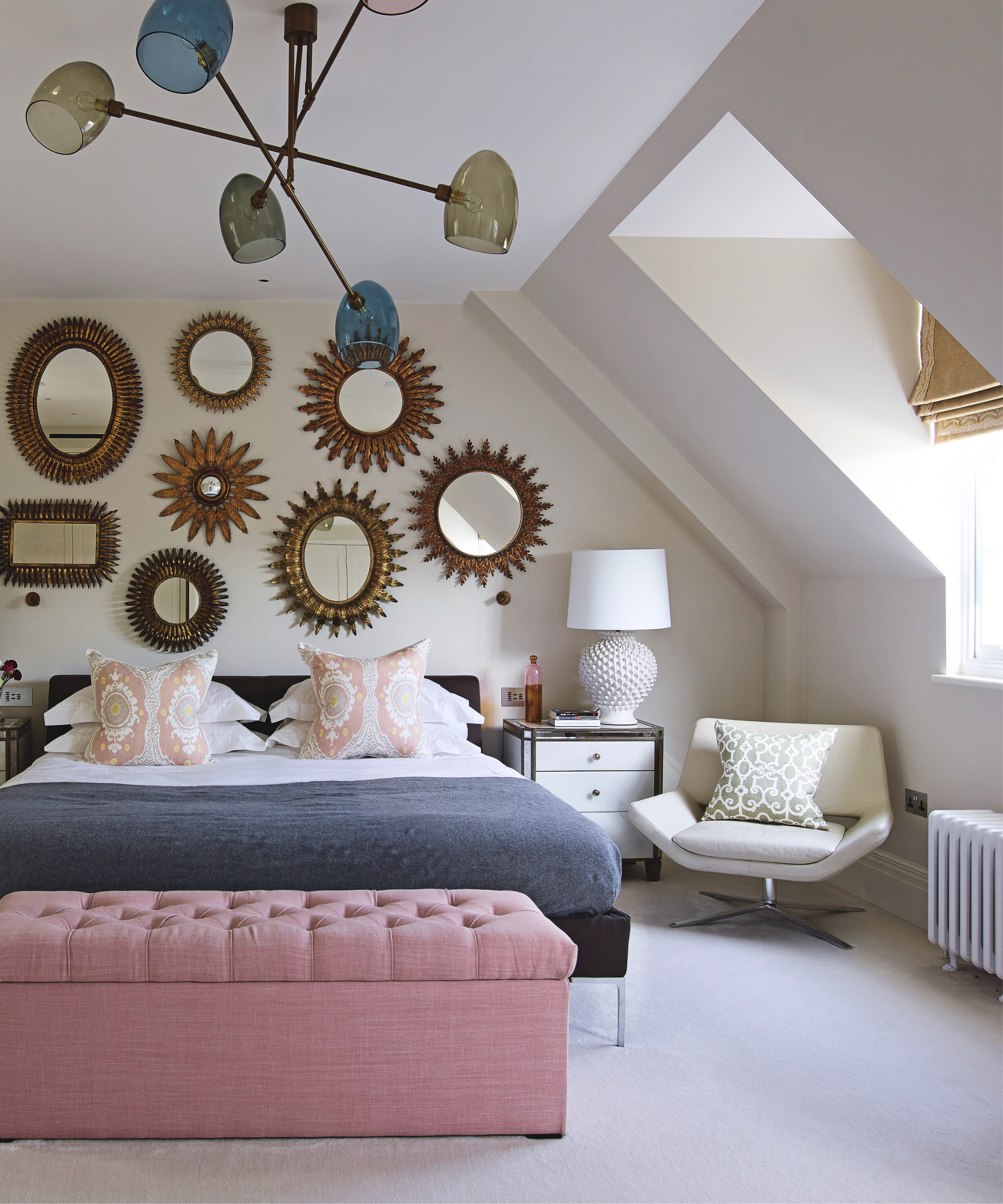 a bedroom with a gallery wall of different sizes of mirrors, a bed with pink, white, and gray bedding, a pink ottoman at the foot of the bed, and an accent chair to the right side