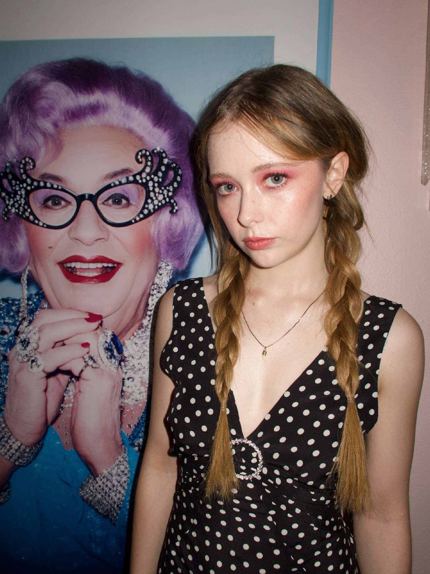 Actress Suzanna Son standing in front of a poster of drag queen. She is wearing a polka dot dress with deep v neck and her hair in braids.