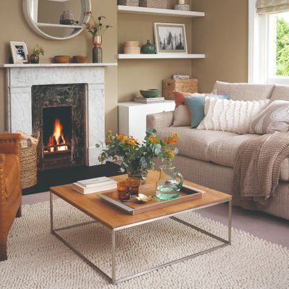 A beige-painted living room with a lit fireplace and a beige sofa with multi-coloured scatter cushions