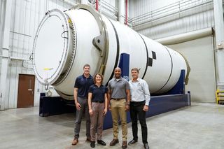 four humans stand in front of a large white cylinder laid on its side.