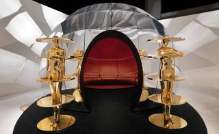 View of Novembre’s room installation - a silver reflective dome with red interior, black flooring that begins outside the dome and two gold female statues with gold discs sitting at the head, bust, hips and knees located by the entrance to the dome