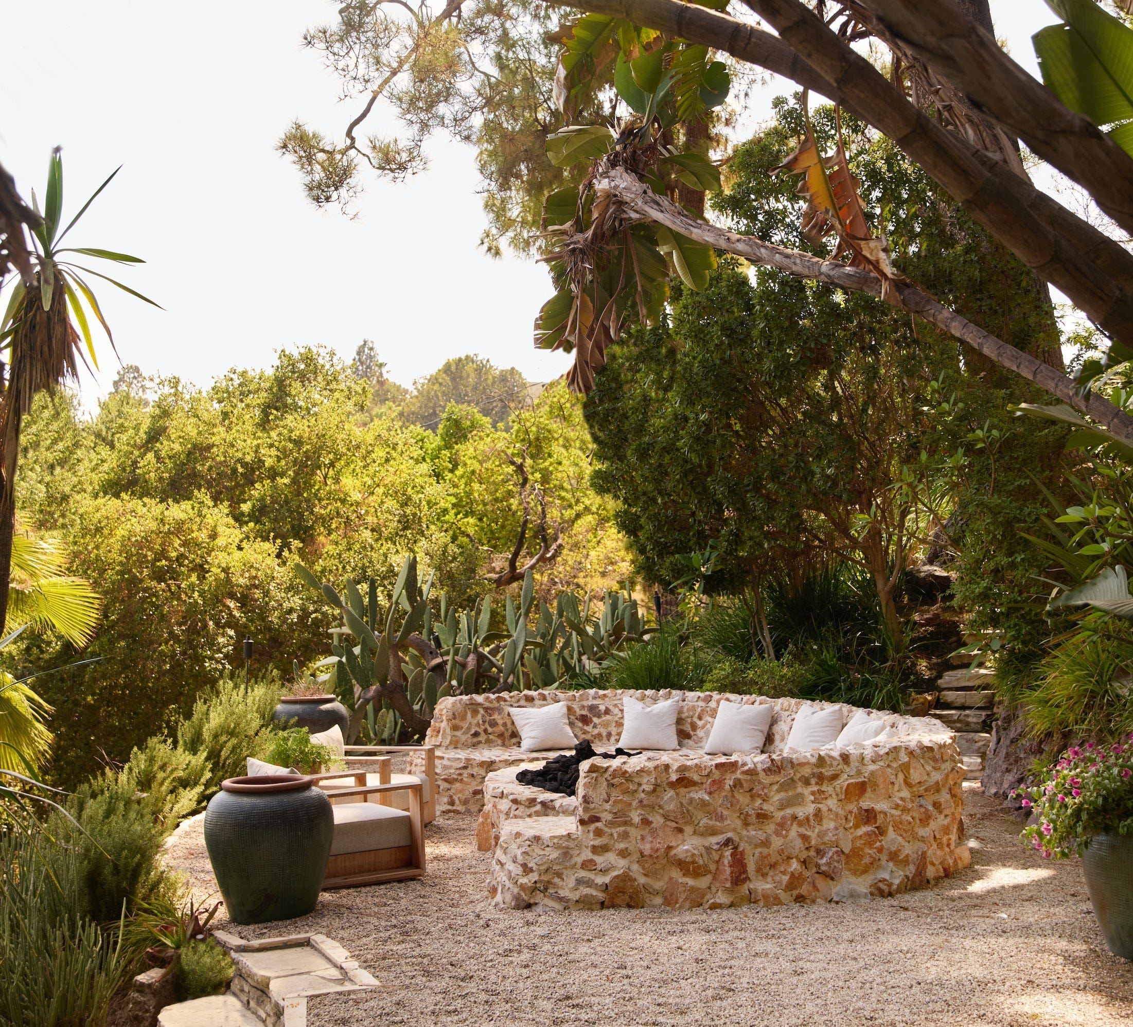stone semi-circle shaped seating around a firepit with white cushions. Two green vases and green trees surround the seating