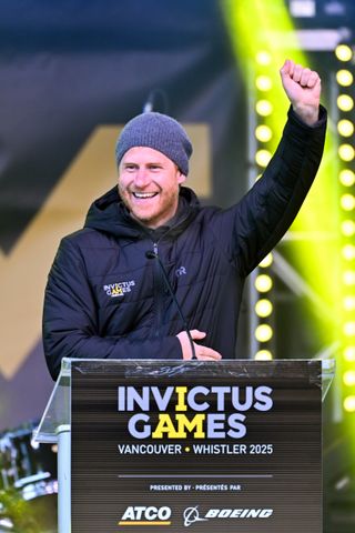 Prince Harry wearing a black Invictus Games coat smiling and raising his arm up at a podium reading Invictus Games Vancouver Whistler 2025