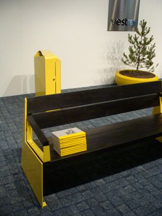 A brown and Yellow outdoor bench with white and yellow books stacked on the bench. Behind the bench is a yellow bin and yellow pot with a tree against a white wall