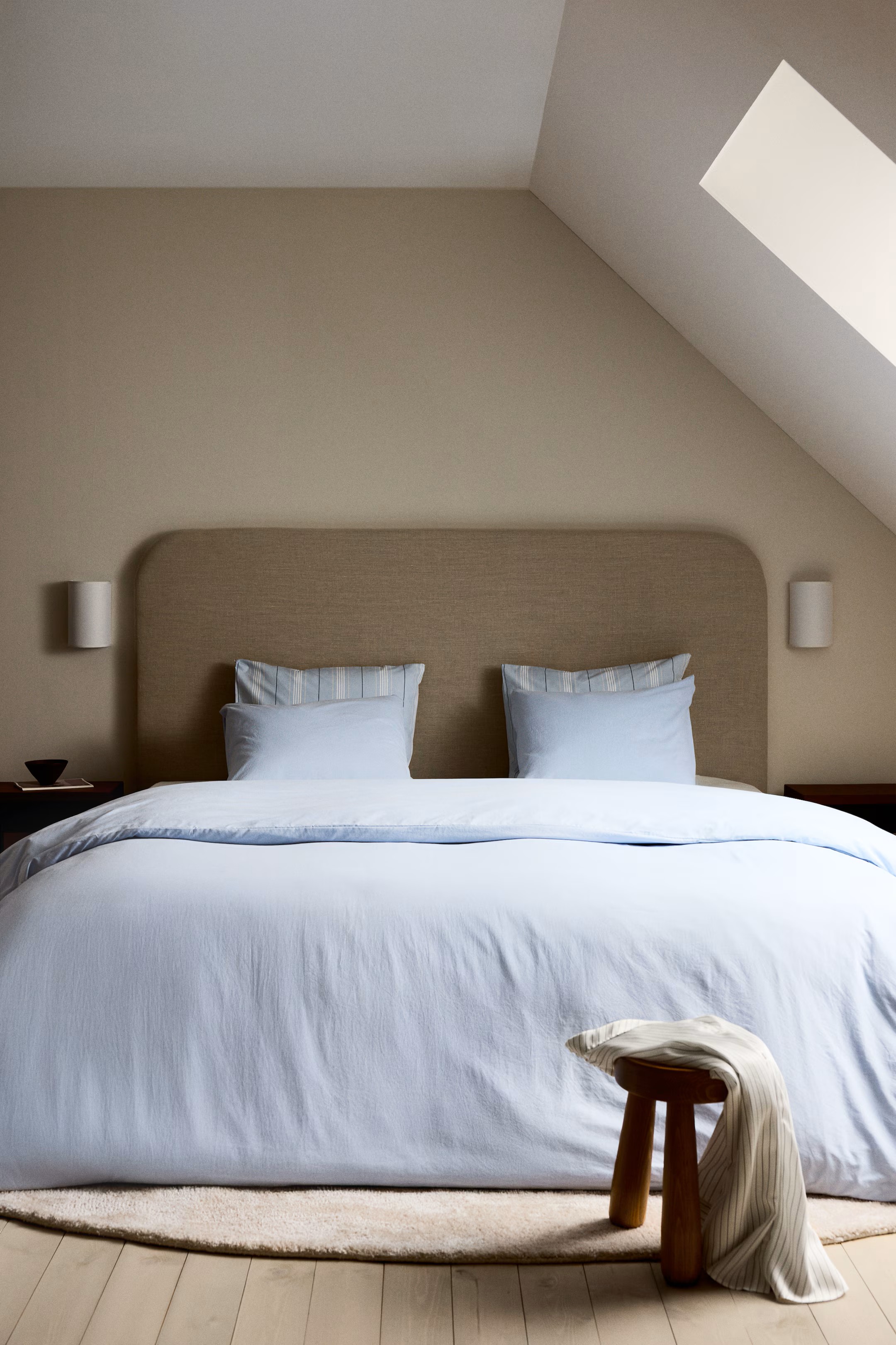 Image of a large bed with light blue bedding and a beige headboard in a minimalist, beige bedroom.