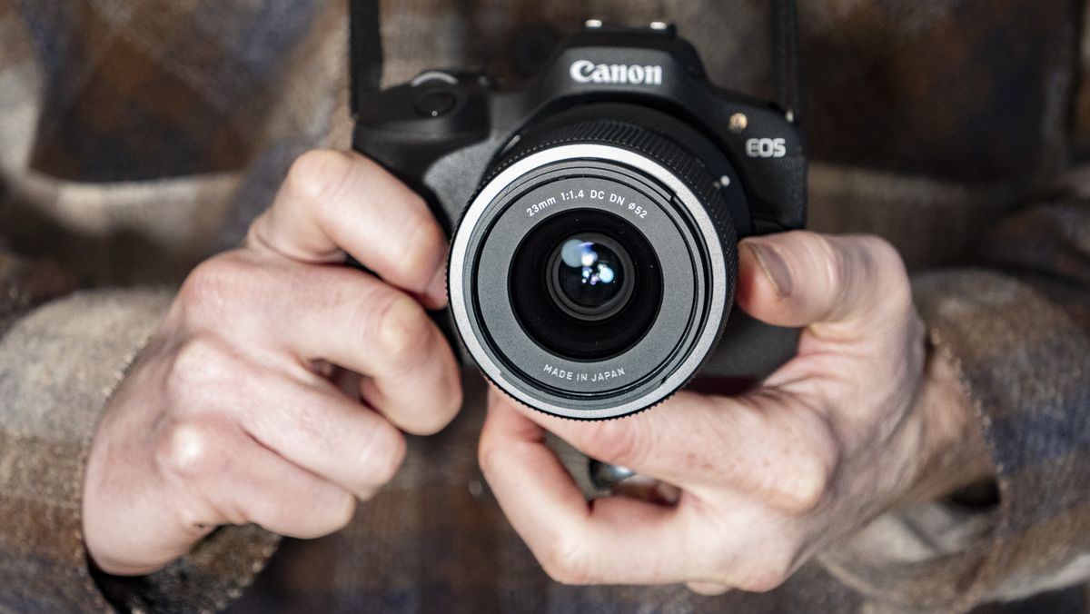 Sigma 23mm F1.4 DC DN Contemporary lens in the hand attached to a Canon EOS R100, checked shirt background