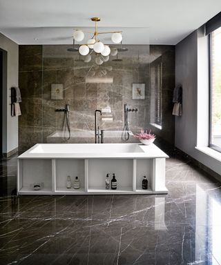 Bathroom with gray marbled floor and walls and large central white bath, large hanging modern chandelier over bath