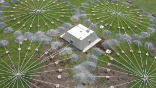 as seen from above, four circles composed of wires radiating out from central points on the ground surround a low square building in a field