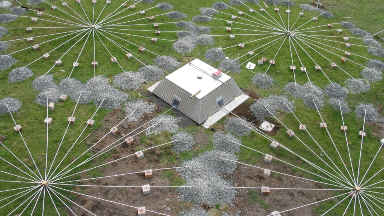 as seen from above, four circles composed of wires radiating out from central points on the ground surround a low square building in a field