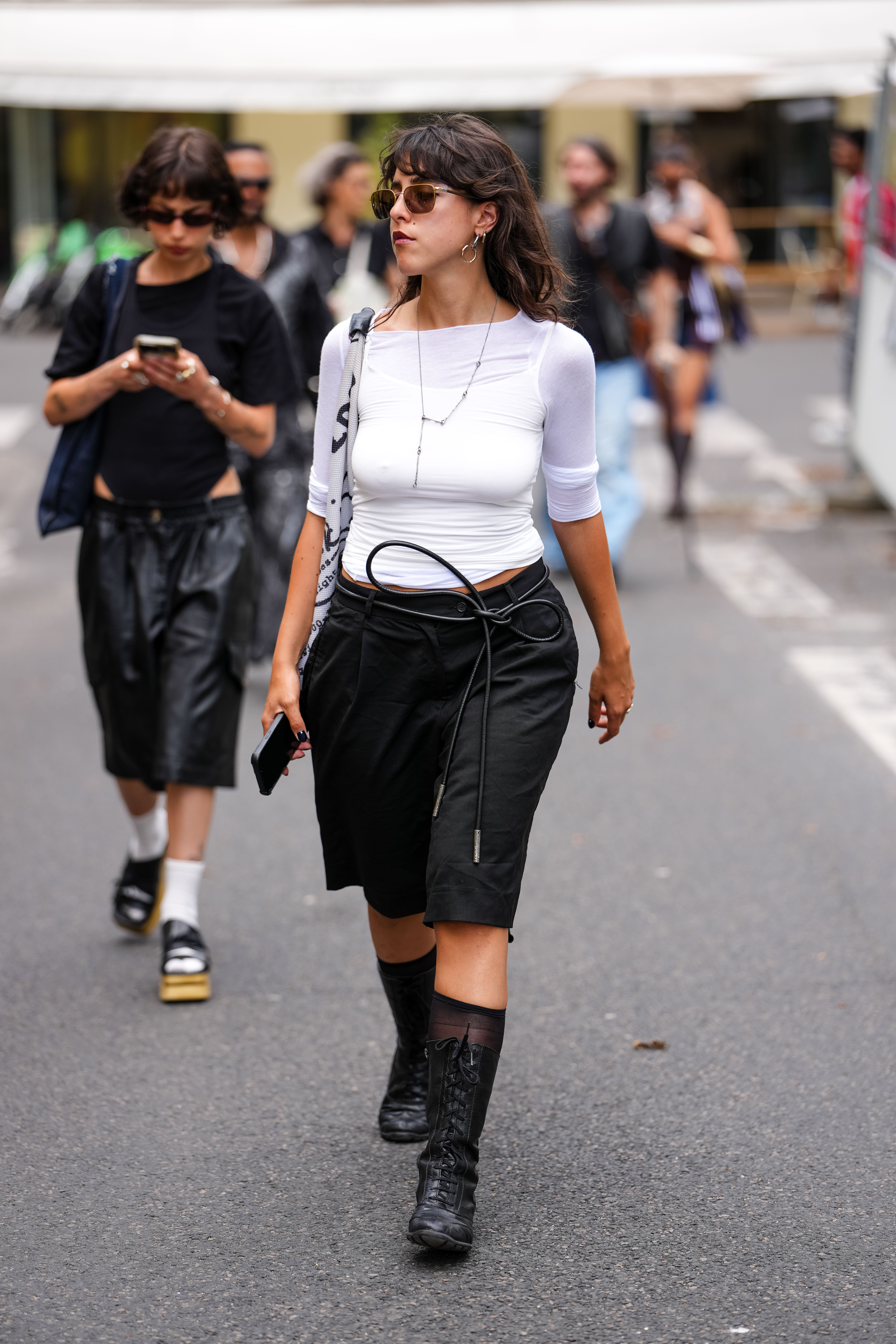 PARIS, FRANCE - JUNE 26: A guest wears a fitted white top with three-quarter sleeves. Sunglasses with a brown tint are worn, along with hoop earrings and a delicate chain necklace. A black crossbody bag with a visible strap is carried. Black knee-length shorts with a drawstring detail are paired with black lace-up boots featuring a mid-calf length. The hair is styled in loose waves, and a smartphone is held in one hand, outside Bluemarble, during Paris Fashion Week - Menswear Spring/Summer 2026, on June 26, 2025 in Paris, France (Photo by Edward Berthelot/Getty Images)