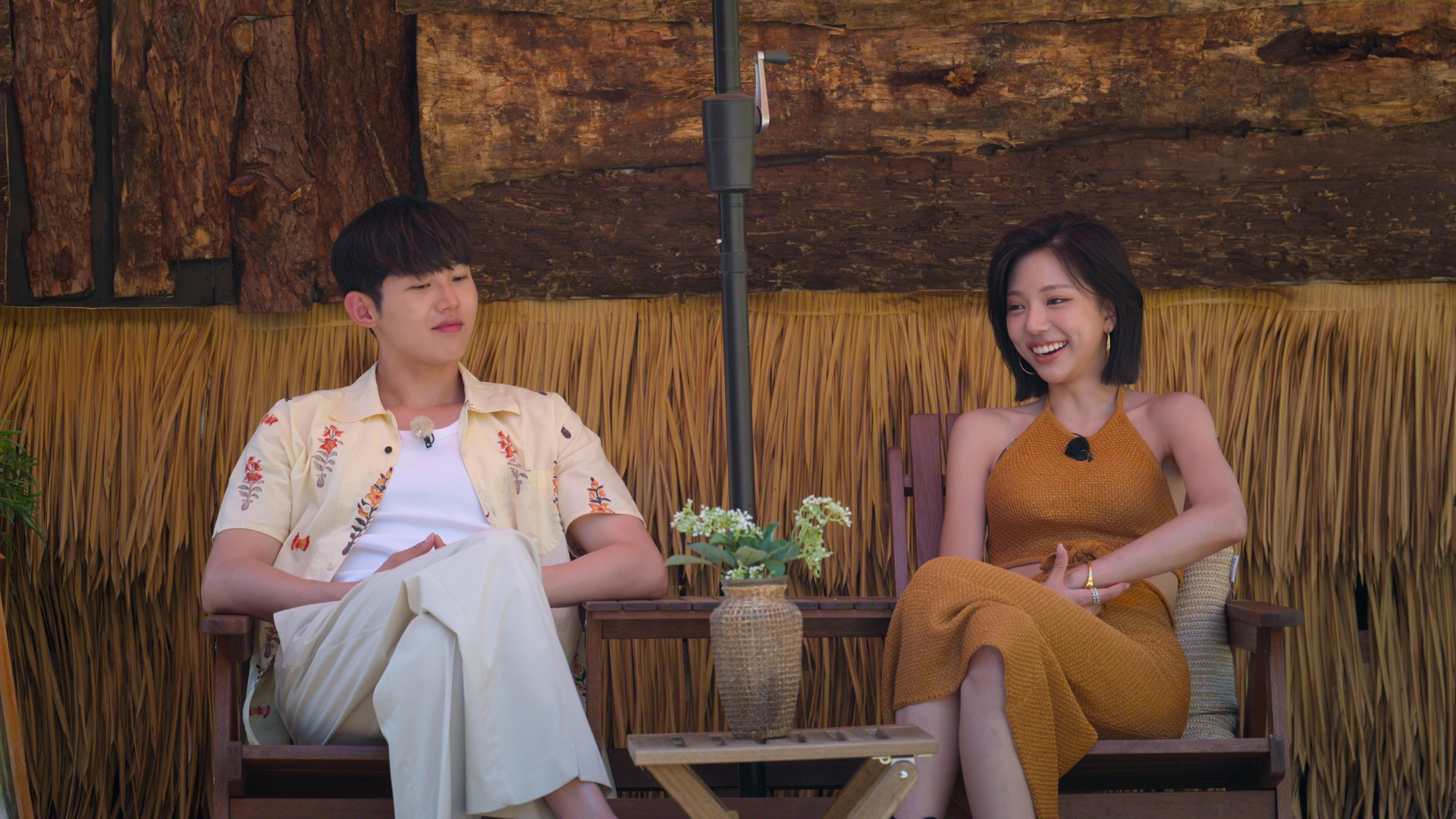 A man (Woo Sung-min) and a woman (Kim Go-eun) chat while sitting at a wooden outdoor furniture set in front of a raffia and wooden structure. The man wears a printed button-down over a white shirt and white pants, while the woman wears a brown halter dress. A still from 'Single's Inferno' season 5.
