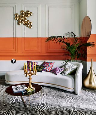 Living room with grey painted walls and paneling, striking orange stripe painted horizontally across walls, grey sofa and zebra print carpet, metallic candle holder and wall art, palm plant