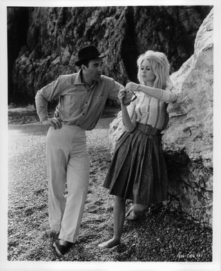 Michel Piccoli talking to Brigitte Bardot as she's playing with sunglasses and wearing a striped henley shirt and A-line skirt in a scene from the film Contempt, 1963.