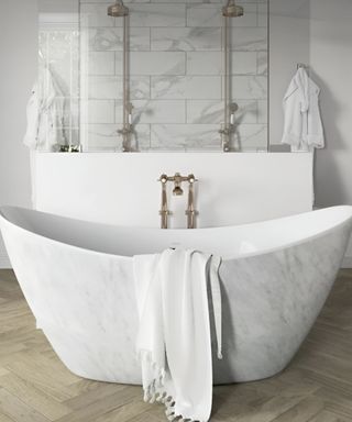 A white and gray marble freestanding bathtub with a white towel in it with a white shelf and two silver shower heads with marble subway tiles behind it