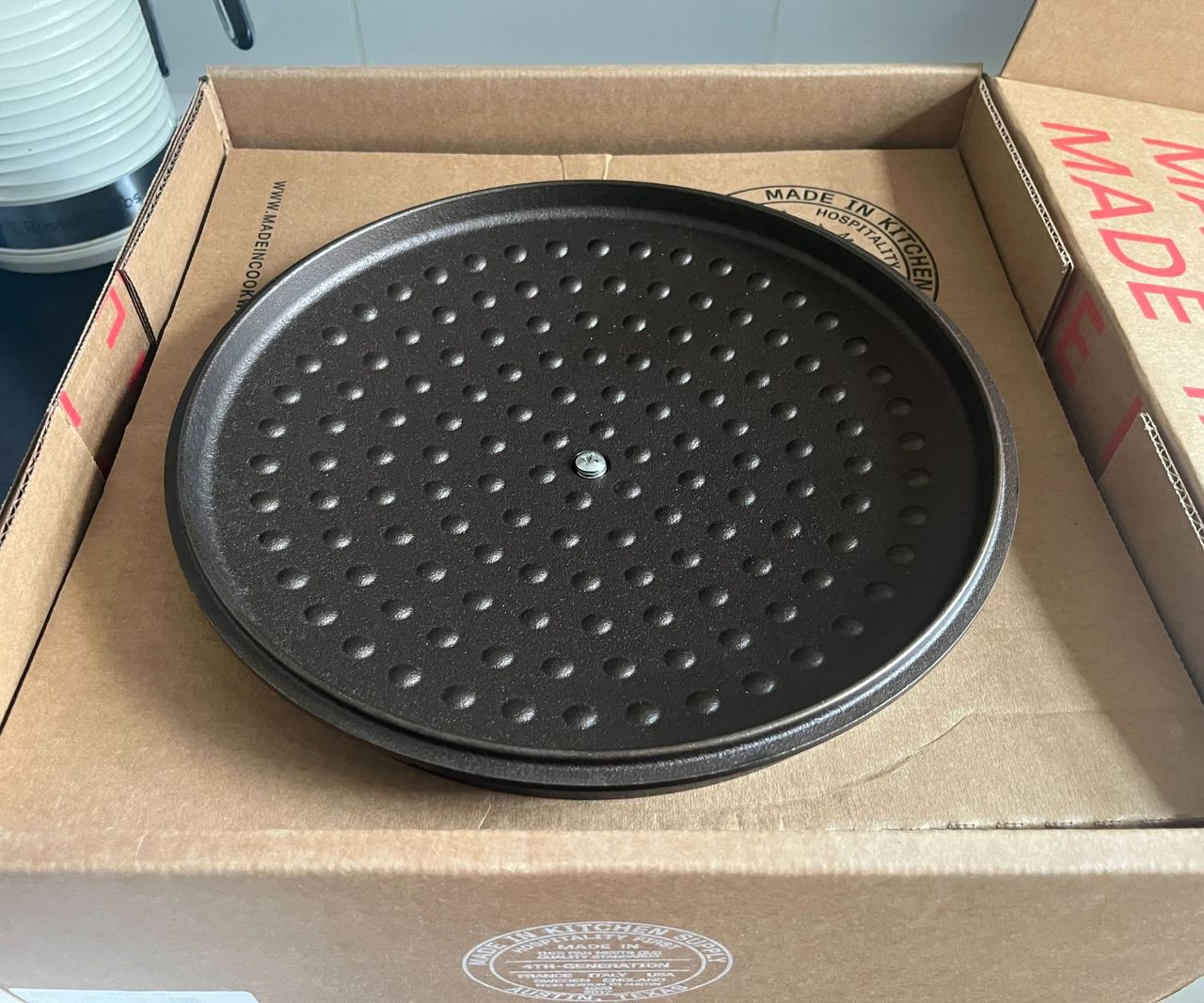 A Made In cookware cardboard box on a black kitchen counter with an upside down Dutch oven lid inside.