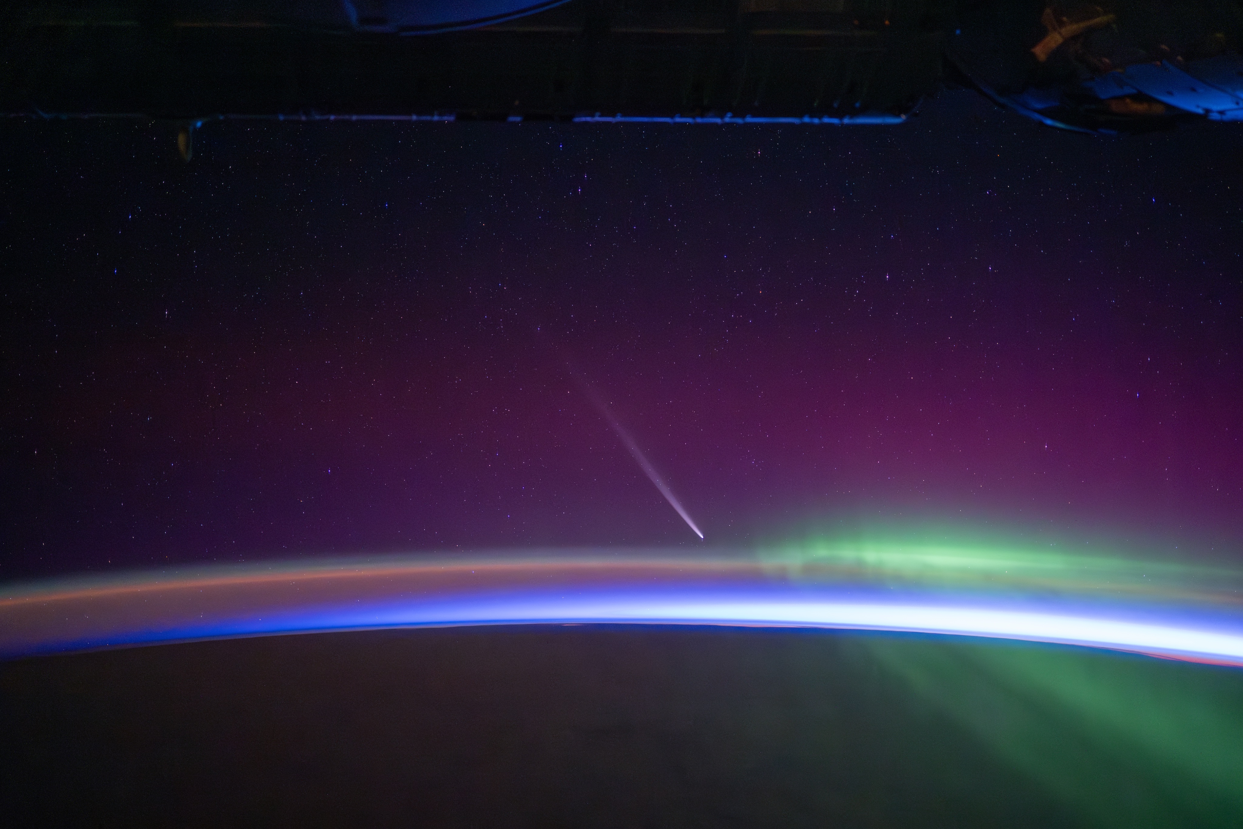 A brilliant comet with a long tail shining over a nighttime Earth and green aurora as seen from space