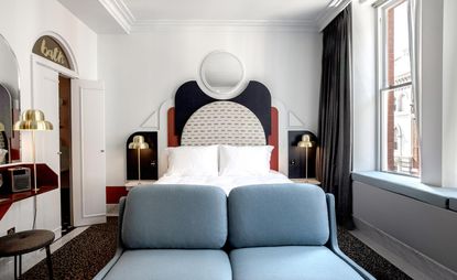 A room in the Henrietta Hotel. A large bed with white linen and geometric headboard in beige, white, red, and black is on the far wall. In front of the bed, there is a sitting place in light blue. White marble tiles are covered with a dark rug. To the right, there is a large window.
