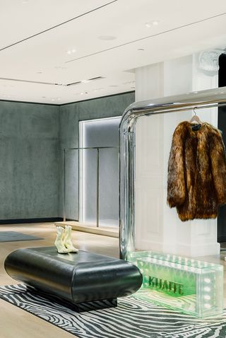 Black stool with a beige pair of heels next to a silver rail with brown fluffy coat hanging